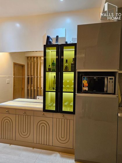 A minimalist crockery and bar unit integrated with the kitchen. The design features a lit glass cabinet for display, a built-in microwave, and handleless base cabinets with a routed arch pattern.
