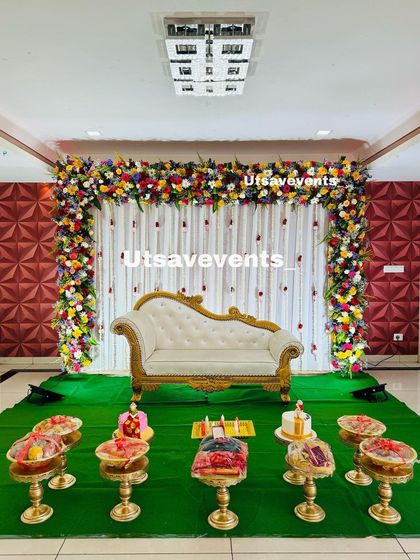 A baby shower stage with a lush arch of colorful fresh flowers over a white chaise lounge. The green carpet and traditional plate settings complete the look.