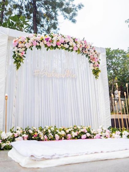 A clean and elegant stage setup for an outdoor engagement. A backdrop of white drapes is accented with a beautiful arch of pink and white roses, flanked by modern gold candle stands.