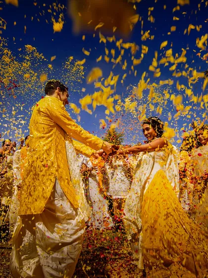 A couple holds hands as they are showered with yellow petals against a brilliant blue sky. This vibrant, colorful shot is the epitome of a joyous haldi celebration.