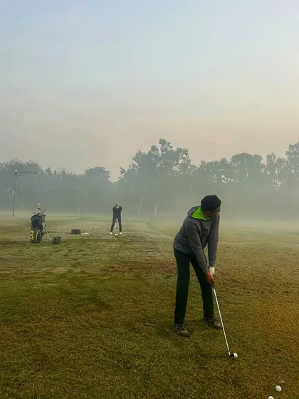 Two golfers practice in the thick morning fog, their silhouettes barely visible against the haze.