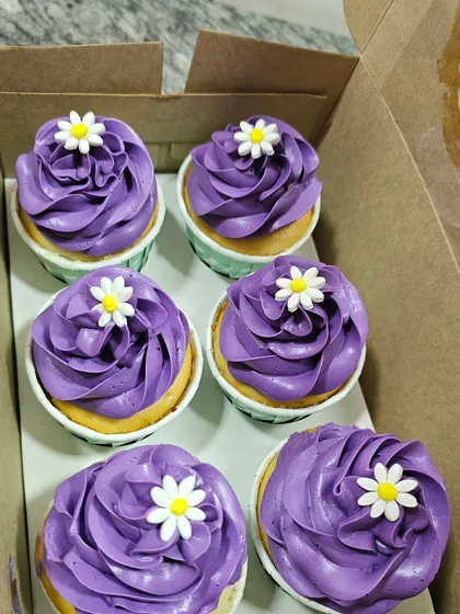 A box of beautiful purple buttercream cupcakes, each topped with a simple, elegant daisy.