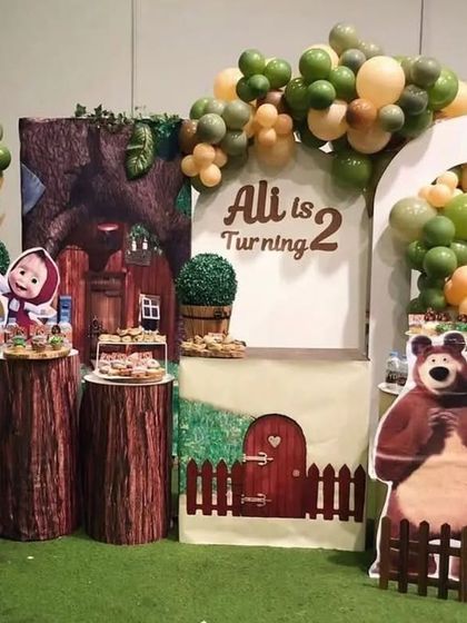 A charming Masha and the Bear theme set in a forest-like environment. The decor includes tree-stump plinths, character cutouts, and a backdrop of Masha's house.