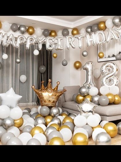 An 18th birthday living room setup. The decor includes "Happy Birthday" foil letters, number balloons, and a floor covered in gold, silver, and white balloons.