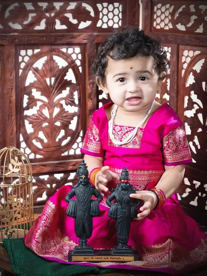 A one-year-old girl in a beautiful pink Paithani-style dress, holding miniature idols of Vitthal and Rakhumai.