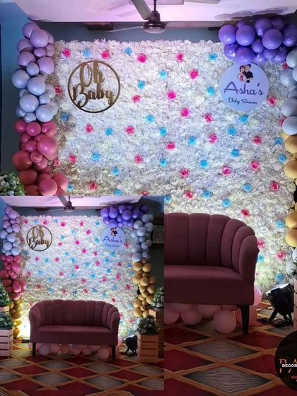 An elegant "Oh Baby" shower backdrop. This design features a wall of white flowers with pops of color, framed by a multi-colored balloon garland, and a custom wooden sign.