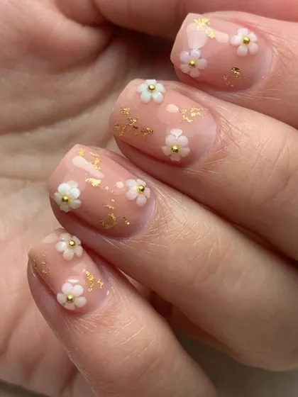 Nail inspiration showing a close-up of short nails with a nude base, gold foil, and tiny 3D white flowers.