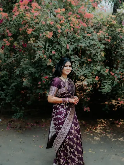 A full-length shot of the bride-to-be, showcasing her elegant purple saree against a natural, floral background.