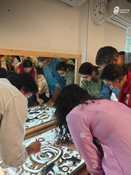 A group of parents and educators gather around a light table, using sand and loose parts to create intricate designs. This collaborative play demonstrates how our learning environment encourages curiosity and connection for all ages.