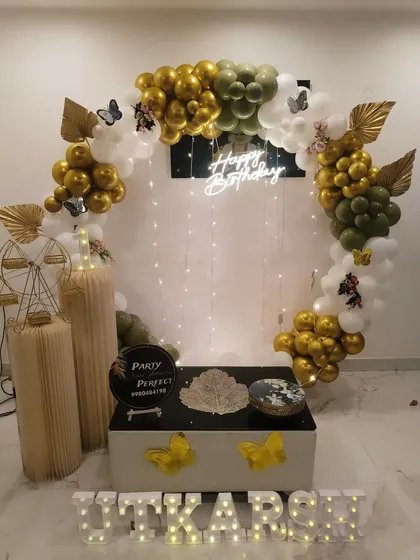 A sophisticated first birthday setup with an olive green, gold, and white balloon arch. The decor includes butterfly accents, a fairy light curtain, a decorative Ferris wheel, and the name 'UTKARSH' in lights.