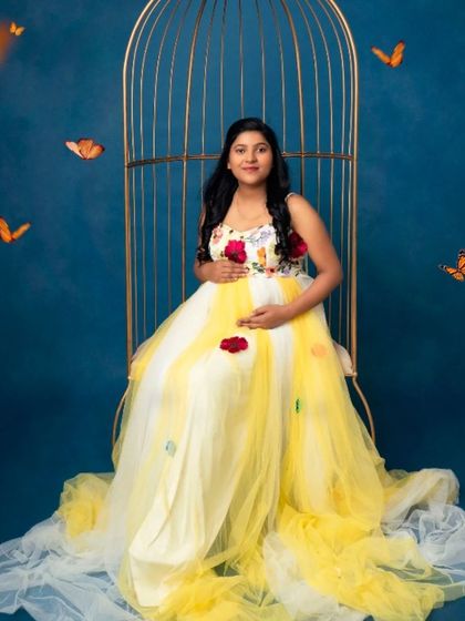 A whimsical portrait of a mom-to-be in our yellow and white floral gown, seated in a golden birdcage surrounded by butterflies.