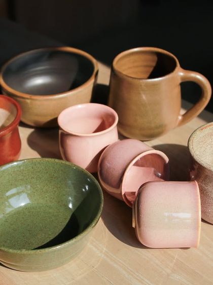 An assortment of cups, bowls, and mugs in earthy green and soft pink glazes, created by students in our 7 week wheel throwing course.