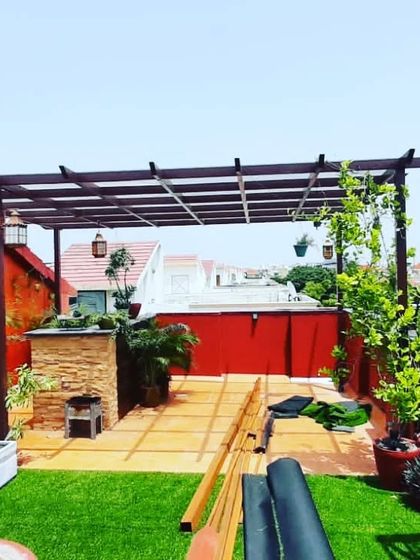 A work-in-progress shot of a large terrace with a barbecue area under a pergola, surrounded by artificial grass and planters.
