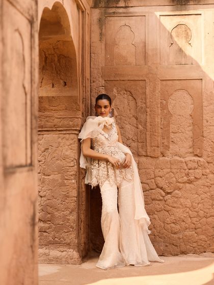 This look features a contemporary sharara with an organza ruffle detail. The styling emphasizes the outfit's fluid silhouette and intricate embellishments, set against a textured, sunlit wall.