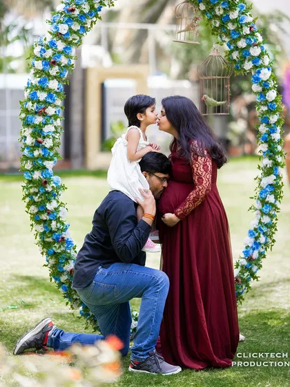 A heartwarming family maternity photo capturing a beautiful moment of connection. The older sibling shares a kiss with mom while dad listens to the baby bump, all framed by a lovely floral arch.