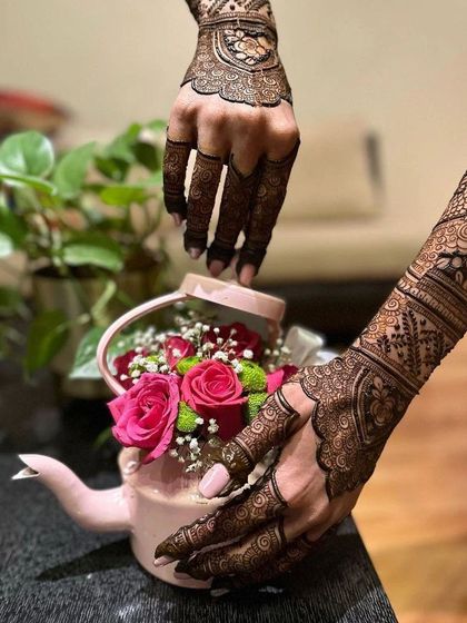 A creative shot of a detailed engagement mehendi. The design features delicate leaf patterns on the fingers and a mix of traditional paisleys and florals on the hand and arm.