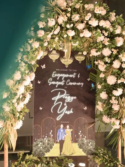 A custom welcome sign for Pragya & Yash's Engagement and Sangeet, framed by a stunning arch of white flowers and greenery.