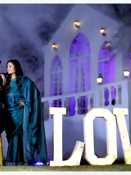 A powerful and romantic pre-wedding portrait. The couple stands confidently in front of the "LOVE" sign, with dramatic smoke and lighting creating a movie-poster feel.