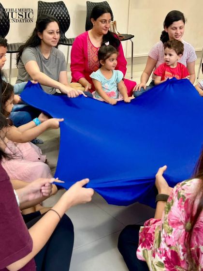 Parents and toddlers play with a large blue cloth, creating waves of music and movement together. This group activity is great for developing social skills and coordination.