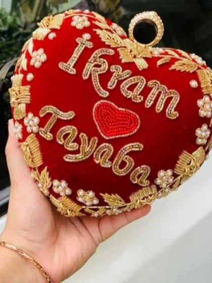 My heart-shaped bridal clutch, made from rich red velvet, is one of my favorite babies. I hand-embroider your names, like 'Ikram' and 'Tayaba' here, to make it a keepsake you'll treasure long after the wedding day. It's perfect for holding your phone and lipstick.