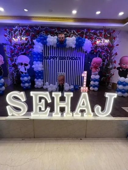 A Boss Baby themed first birthday stage for Sehaj. This large-scale decoration includes a striped backdrop, multiple character cutouts on balloon pillars, and giant light-up letters for the name.