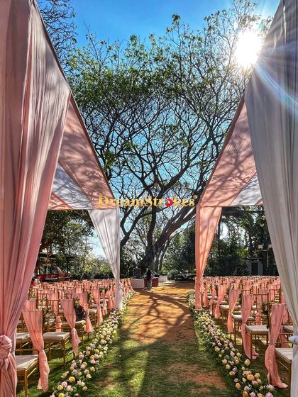 A sun-drenched view of the wedding aisle, lined with Tiffany chairs and pastel peach drapes, creating a dreamy and romantic pathway for the bride.