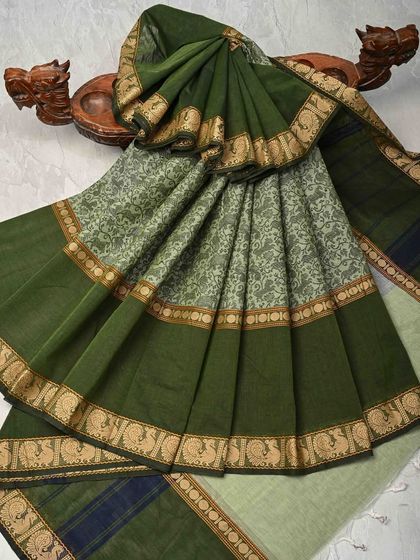 An olive green cotton saree with a printed body and a traditional peacock border. The earthy color and comfortable fabric make it a great choice for daily wear.