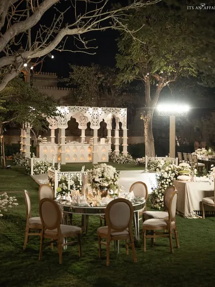 An elegant 'Sheesh Mahal' themed reception at The Oberoi Udaivilas. Mirrored tables reflect the candlelight and intricate white structures, creating a sophisticated and luxurious outdoor dining experience under the stars.