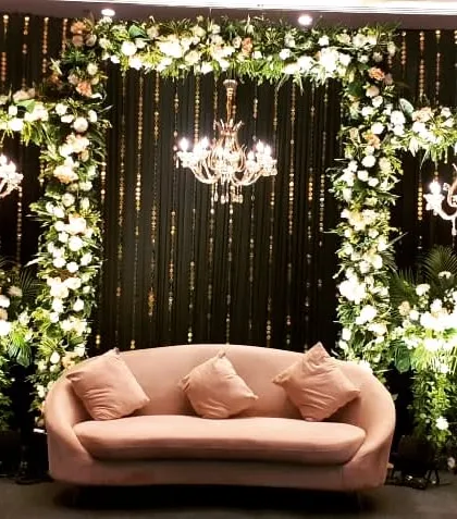 An elegant engagement or reception stage with a dark backdrop that makes the white floral arches and chandeliers pop. The simple pink sofa adds a soft touch of color.