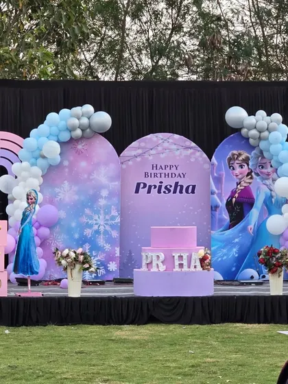 For this 'Frozen' themed birthday, I designed a magical winter wonderland stage. The setup includes custom printed arched panels featuring Elsa and Anna, snowflake details, and a color-coordinated balloon garland in shades of blue, white, and lavender.