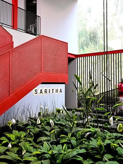 This image captures the relationship between the striking red staircase and the indoor garden at the SARITHA SERENE office. The design juxtaposes hard, industrial geometry with soft, organic plant life.