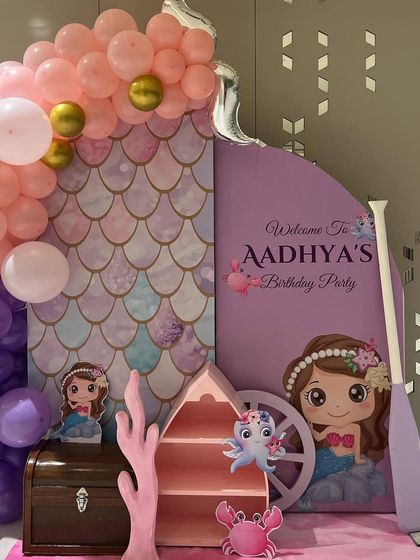 A magical entrance for a mermaid-themed birthday party. The decor features a mermaid-scale patterned wall, a personalized welcome sign, and a beautiful arch of pink and purple balloons.