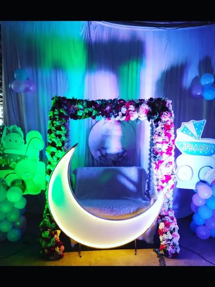 A baby shower decoration with a lighted moon prop and a floral swing. The 'It's a Boy' and 'It's a Girl' cutouts make it perfect for a gender-neutral or reveal party.