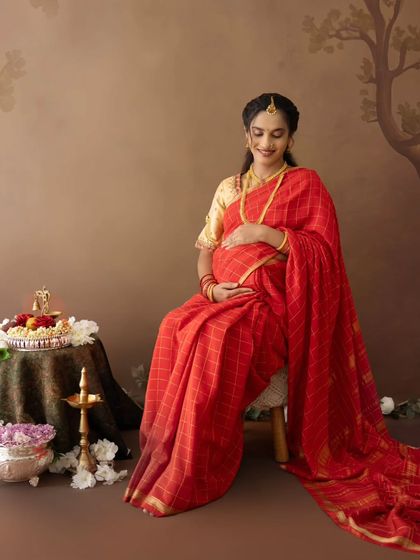 A serene portrait of a mom-to-be in a red checked saree, seated beside a traditional arrangement of flowers and a diya, perfect for a baby shower photoshoot.