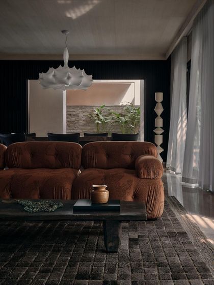 Sunlight streams into the living room, highlighting the rich texture of the sofa and the dark, patterned rug. The space is designed to feel comfortable and lived-in.