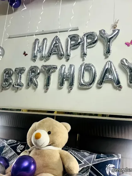 Another close-up of the birthday setup, focusing on the 'Happy Birthday' banner and the cute teddy bear. The silver and purple color scheme creates a modern and elegant look.