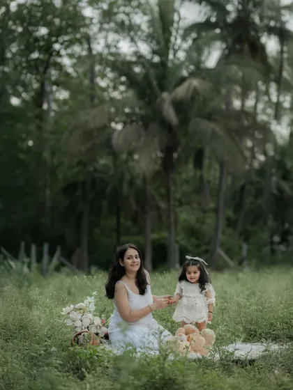 A mother and daughter enjoy a picnic setup in a beautiful meadow surrounded by tall trees. The mother holds her daughter's hand, sharing a sweet moment of connection amidst the beauty of the outdoors.