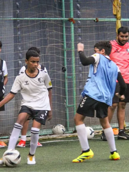 A player shields the ball from a defender under the watchful eye of a coach, applying game-like pressure in a training scenario.