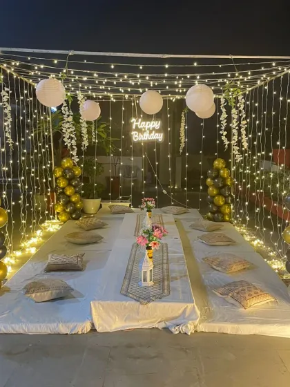 We created this cozy and intimate birthday celebration on a terrace. The setup features a canopy of fairy lights, low floor seating with comfortable cushions, and elegant black and gold balloon columns, perfect for a memorable evening under the stars.