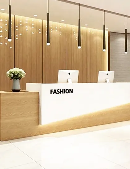 A clean and modern reception desk combining light wood with a crisp white counter. The backlit wood-paneled wall with a geometric pattern and hanging pendant lights adds a touch of creative flair to the office space.