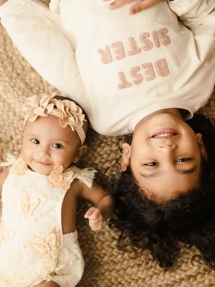 An adorable overhead shot of two sisters lying on a jute rug. The older sister's "Best Sister" shirt is a cute detail in this playful sibling portrait.