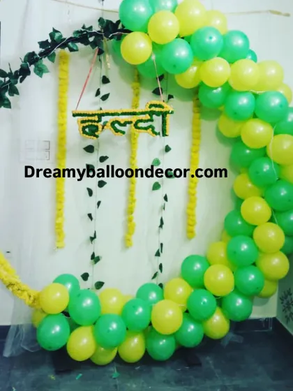 For a Haldi ceremony, I can create a beautiful and modern photo booth. This setup uses a circular frame decorated with a balloon arch in traditional Haldi colors and fresh marigold garlands, with a hanging 'Haldi' sign in Hindi.