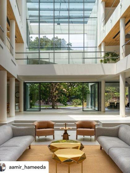 A grand, double-height living space with minimalist furniture that opens directly onto a view of the garden and pool. The architecture is designed to maximize the connection to the outdoors, creating a sense of immense space and light.