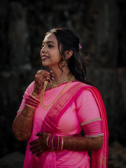 A side-profile portrait of the bride against a dark, textured background. The lighting beautifully accentuates her features, her pink saree, and the intricate henna design.