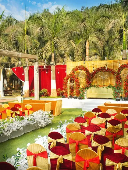 An outdoor wedding ceremony setup with a vibrant red and gold theme. The seating is arranged to give every guest a perfect view of the beautiful floral mandap.