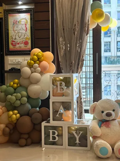 A cozy corner setup for a baby welcome, featuring an earthy-toned balloon garland, a large teddy bear, and 'BABY' blocks.