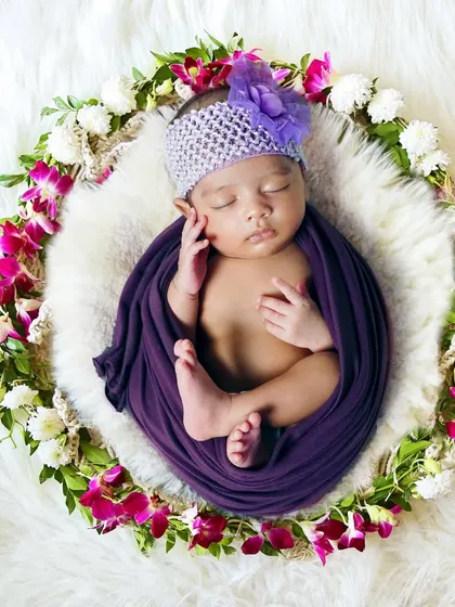 A newborn wrapped in a purple cloth, sleeping in a nest of white fur and a floral wreath.
