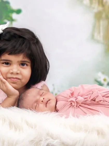 A beautiful portrait of an older sister posing thoughtfully next to her sleeping newborn sibling on a soft, white fur rug.