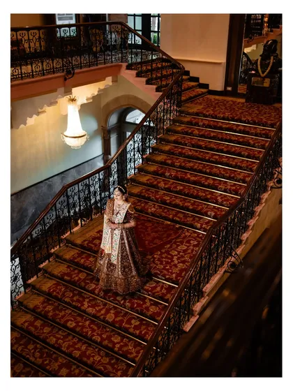 A stunning high-angle shot of the bride descending the grand staircase, her lehenga creating a beautiful trail on the rich red carpet.