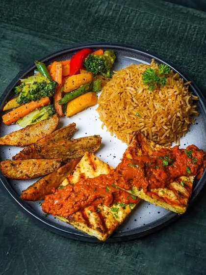 A platter featuring grilled paneer in a red sauce, served with rice, potato wedges, and vegetables. The dark background makes the colors of the meal pop.